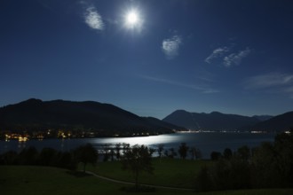 Night scene with moonlight over a calm lake, surrounded by mountain silhouettes and shining lights