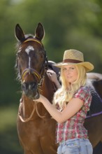 Woman (cowgirl) in a straw hat and plaid shirt stands beside a polish arabian horse on a field,