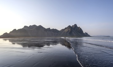 Tourist at black lava beach, sandy beach and sea, mountains Klifatindur reflected in the water,