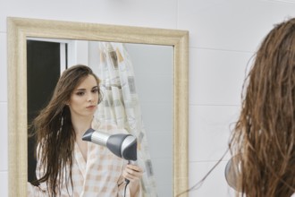 Young woman wearing bathrobe blow drying long hair after shower and looking in mirror, good-looking