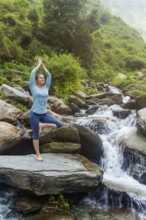 Woman in Hatha yoga balance yoga asana Vrikshasana tree pose at waterfall outdoors