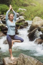 Woman in Hatha yoga balance yoga asana Vrikshasana tree pose at waterfall outdoors