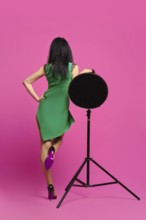 Rear view of woman with black hair and in green dress rising one leg and leaning on studio strobe