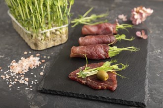 Slices of smoked salted meat with green pea microgreen on black concrete background. Side view,