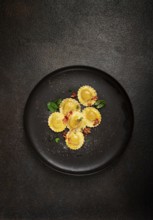 Fresh ravioli, with butter, spices and herbs, fried bacon, on a black plate, top view, dark