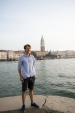 Young man in striped shirt and shorts on the banks of the Grand Canal, behind Campanile, Venice,