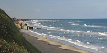 View of the beach with gentle waves lapping against the shore, overgrown with grass and sand dunes,