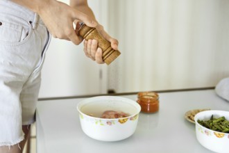 In a bright kitchen, an unrecognizable woman is grinding fresh pepper over chicken pieces in a