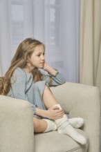 A young pensive girl sits in profile on a soft armchair in a well-lit room with her hand resting