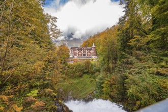 A large hotel surrounded by autumnal trees with mountains and clouds in the background, Lake