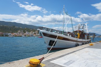 A fishing boat moored on a sunny pier overlooking picturesque hills and coastal houses, Poros,