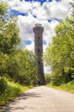 A tall stone tower rises above a road, flanked by trees under a sky full of clouds and sunbeams,