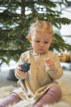 Toddler in a beige dress holding a Christmas tree bauble in front of a festively decorated tree