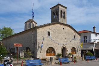 Historic church with bell towers and Greek flags, surrounded by blue benches under a blue sky,