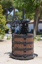 Historische Weinpresse aus Holz und Metall in einem sonnigen Garten mit Bäumen und einem Brunnen,