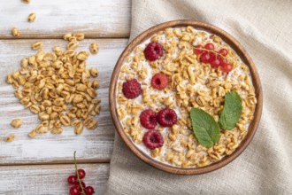 Wheat flakes porridge with milk, raspberry and currant in wooden bowl on white wooden background