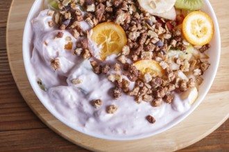 Vegetarian salad of bananas, apples, pears, kumquats, kiwi with granola and yogurt on brown woodem