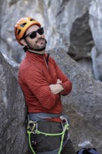 A confident sunglassed climber in orange helmet and red jacket rests against a rock. He smiles with