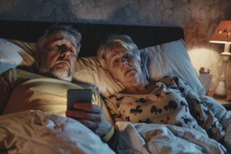 An elderly couple lies in their bedroom in their marital bed and looks at a smartphone, worried,