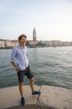 Young man in striped shirt and shorts on the banks of the Grand Canal, behind Campanile, Venice,