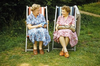 Two women friends sitting in deckchairs on graß lawn one knitting, smiling at each other, England,