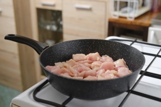 Diced pieces of raw chicken are sizzling in a frying pan on a stovetop. The kitchen has wooden