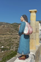 Wooden statue of a woman in traditional dress in front of a mountainous landscape, Olymbos,