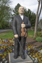 Statue of a man in a dark suit holding a violin stands in a flowering garden under a clear sky,