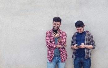 Two smiling friends leaning on a wall checking their cell phones, Friends leaning on a wall texting