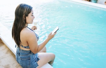 Girl sitting on the swimming pool holding drink and checking cell phone. Smiling girl on vacation