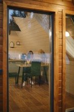 View through the large window at a pensive woman sitting at a table in tiny house in the evening