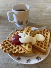 Waffles with jam, cream and cheese on a plate, next to a cup of coffee, served on a wooden table,