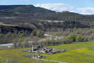 Ruins of an old building in a green field surrounded by a river and wooded hills on a clear spring