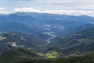 Weitreichende Berglandschaft mit einem grünen Tal, bewaldeten Hängen und einem blauen Himmel mit