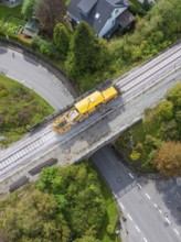 A yellow construction machine moves on railway tracks over a bridge with a road underneath, tamping