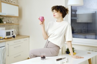 Dreamy and satisfied young woman sits on a kitchen table and holds a glass of smoothie in hand