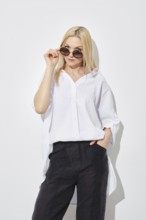 Blonde woman in white shirt and black trousers looking to the side with her sunglasses lowered