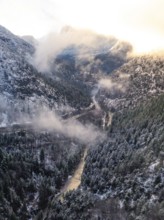 A valley with a river flowing through it surrounded by snow-covered mountains and wafts of mist,