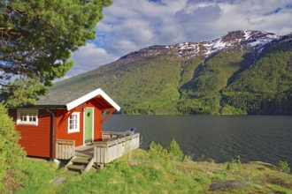 Red-painted cabin on the shore of a fjord against a backdrop of mountains and forest, holiday