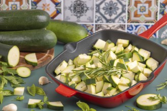 Diced courgettes in a red pan, surrounded by fresh herbs and whole courgettes in front of colourful