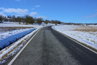 An empty country road leads through a wintry landscape with snow-covered trees and blue sky,