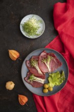 Slices of smoked salted meat with cilantro microgreen on black concrete background and red textile.