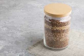 Glass jar with buckwheat poured in layers on a gray concrete background. side view, close up, copy