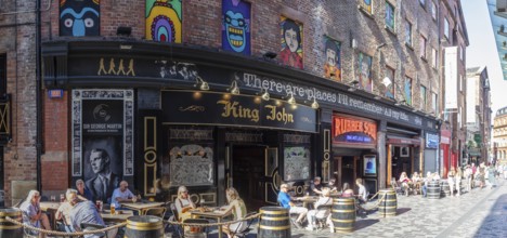 People sitting at tables in front of colourful buildings in a busy street, Liverpool