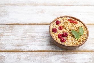 Wheat flakes porridge with milk, raspberry and currant in wooden bowl on white wooden background.