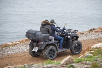 Two people ride a quad bike along a narrow gravel track near the coast, fjord, remote Arctic Inuit