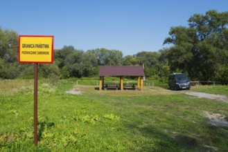 Border sign next to a wooden shelter on a green field with trees and car, motorhome, camper, border