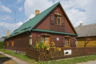 Dark brown wooden house with green roof and flowers in the garden behind the fence, Bialowieza,