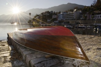 Wood Boat Lying Upside Down on the Waterfront to Lake Maggiore with Mountain in Sunset with Sunbeam