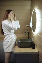 Unrecognizable woman smoothes a sheet mask on her face while standing in the bathroom in front of a